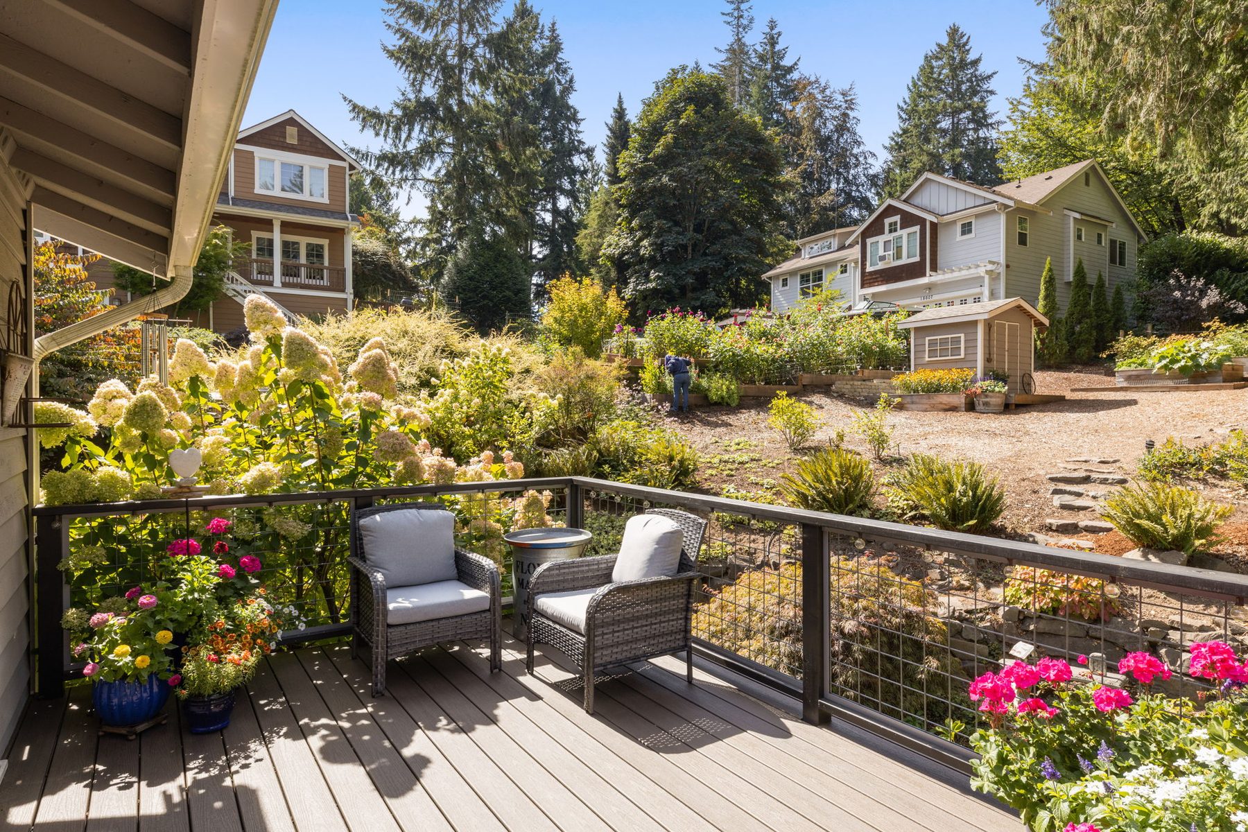 The composite deck with cable railing: two woven armchairs and a low table, planters in flower along the rail, terraced garden beyond.